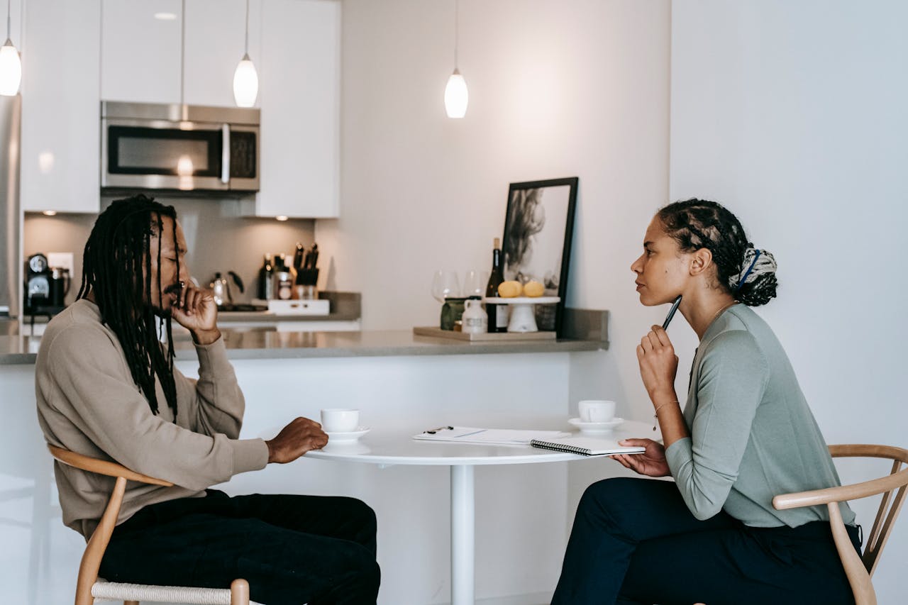Two people sit at a kitchen table having a serious discussion over coffee with a notebook between them.