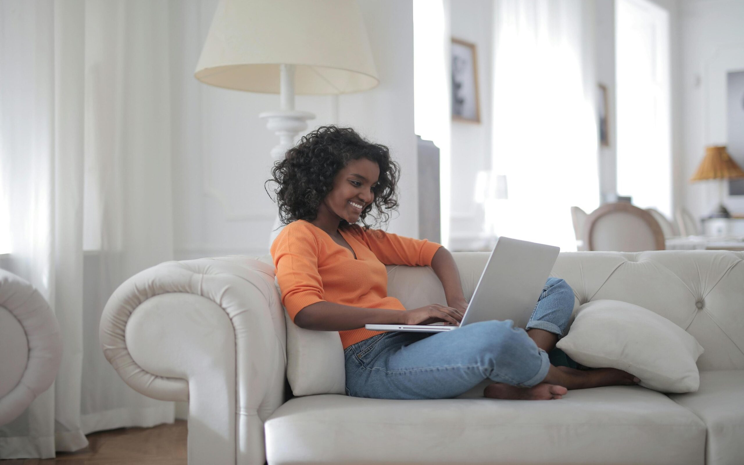 A smiling woman in an orange top works on her laptop while relaxing on a white couch.